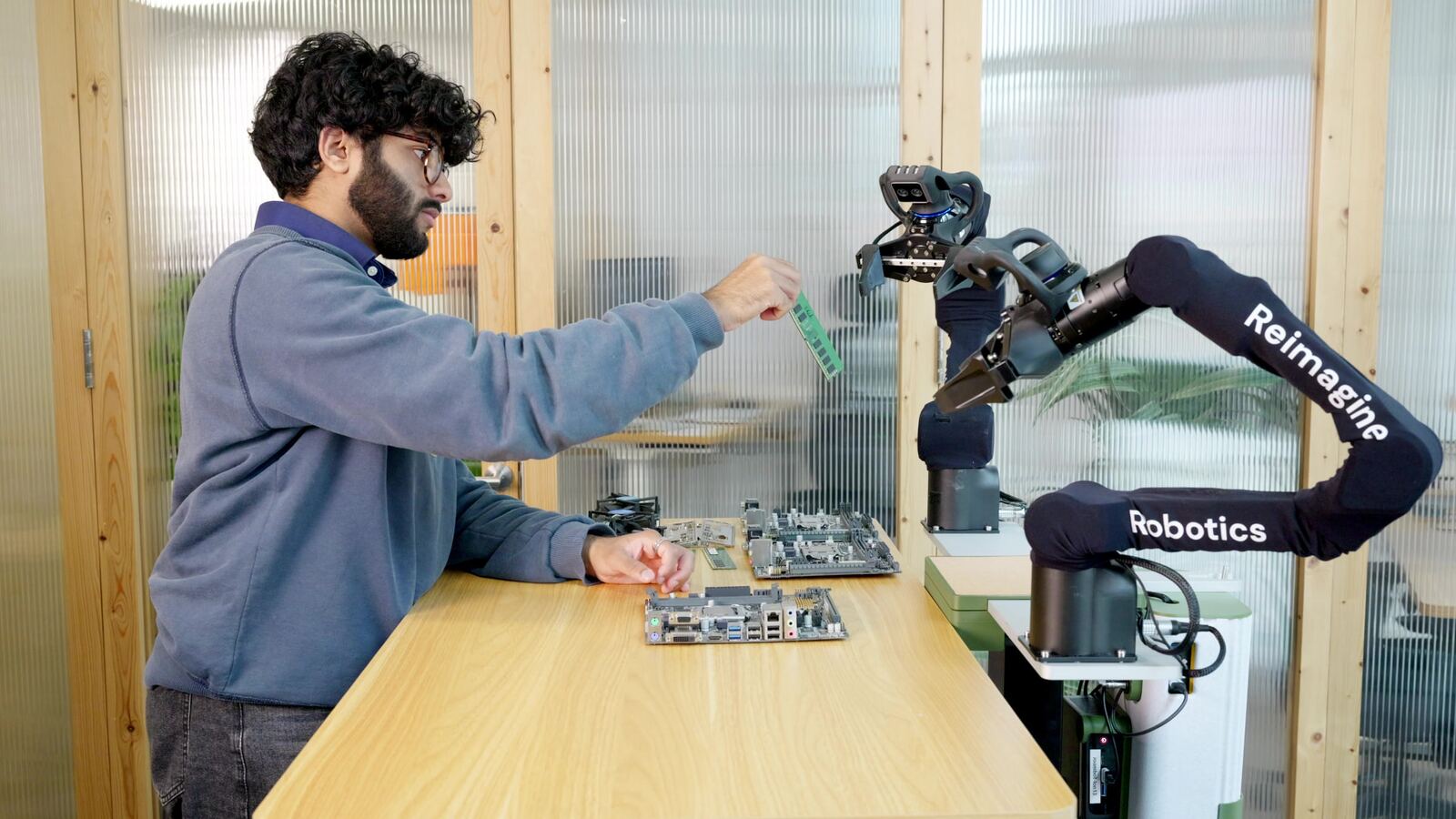 A worker demonstrates a task to a two-armed Reimagine Robotics robot at a workbench.